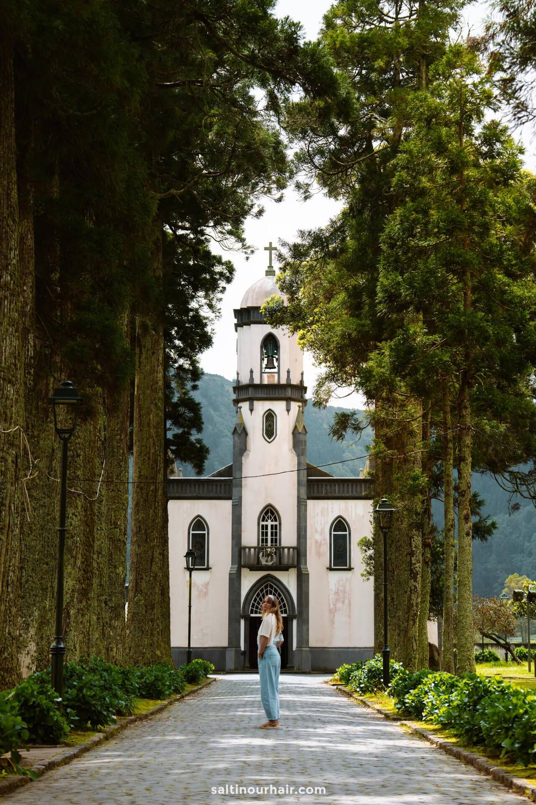A woman in a light blue trousers stands on a tree-lined path facing a white church with a bell tower and cross in Sete Cidades, capturing one of the must-see things to do in S&atilde;o Miguel, Azores.