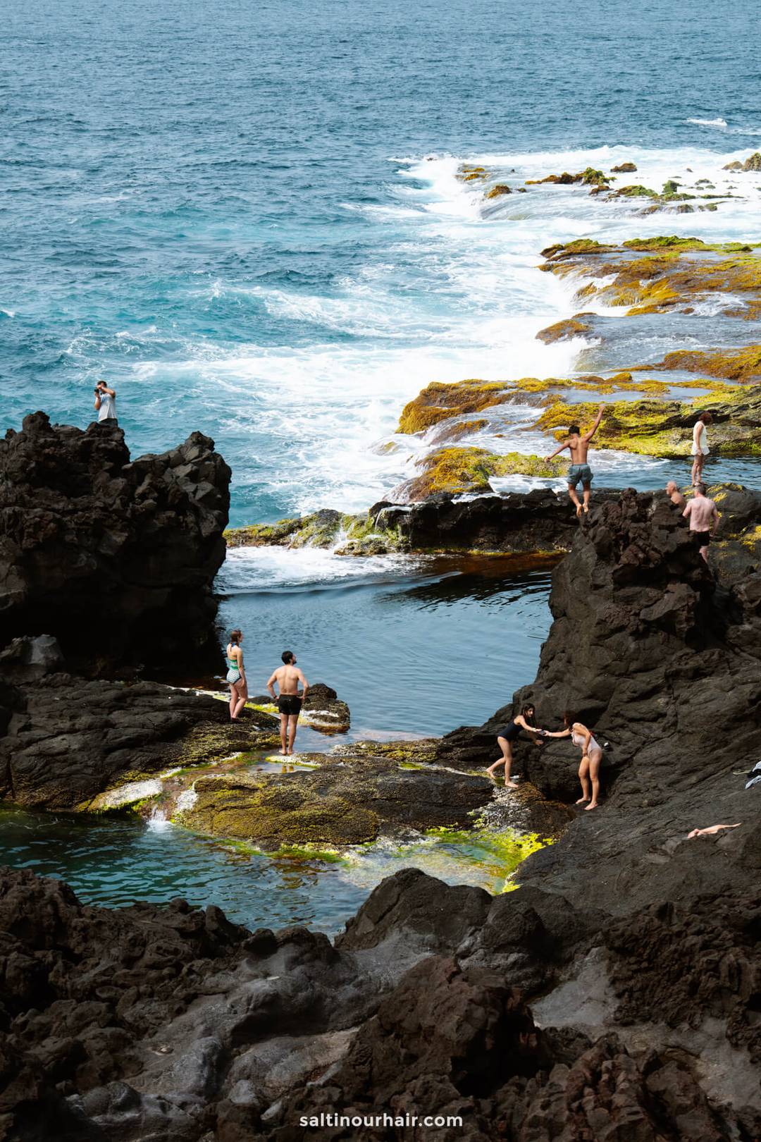 People swim, climb, and relax on rugged black volcanic rocks and tide pools beside the ocean, with waves crashing on the mossy shoreline&mdash;one of the unforgettable things to do in S&atilde;o Miguel, Azores.
