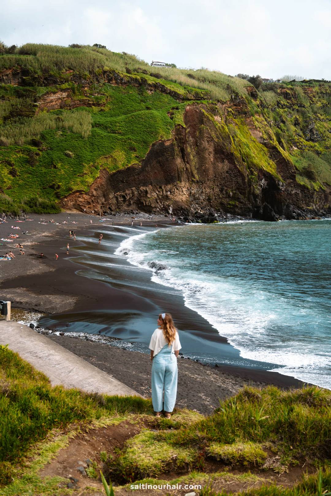 A woman stands on a grassy cliff overlooking a black sand beach called: Praia dos Mosteiros, with people sunbathing and swimming, steep green hills in the background&mdash;a scenic spot among the top things to do in S&atilde;o Miguel, Azores