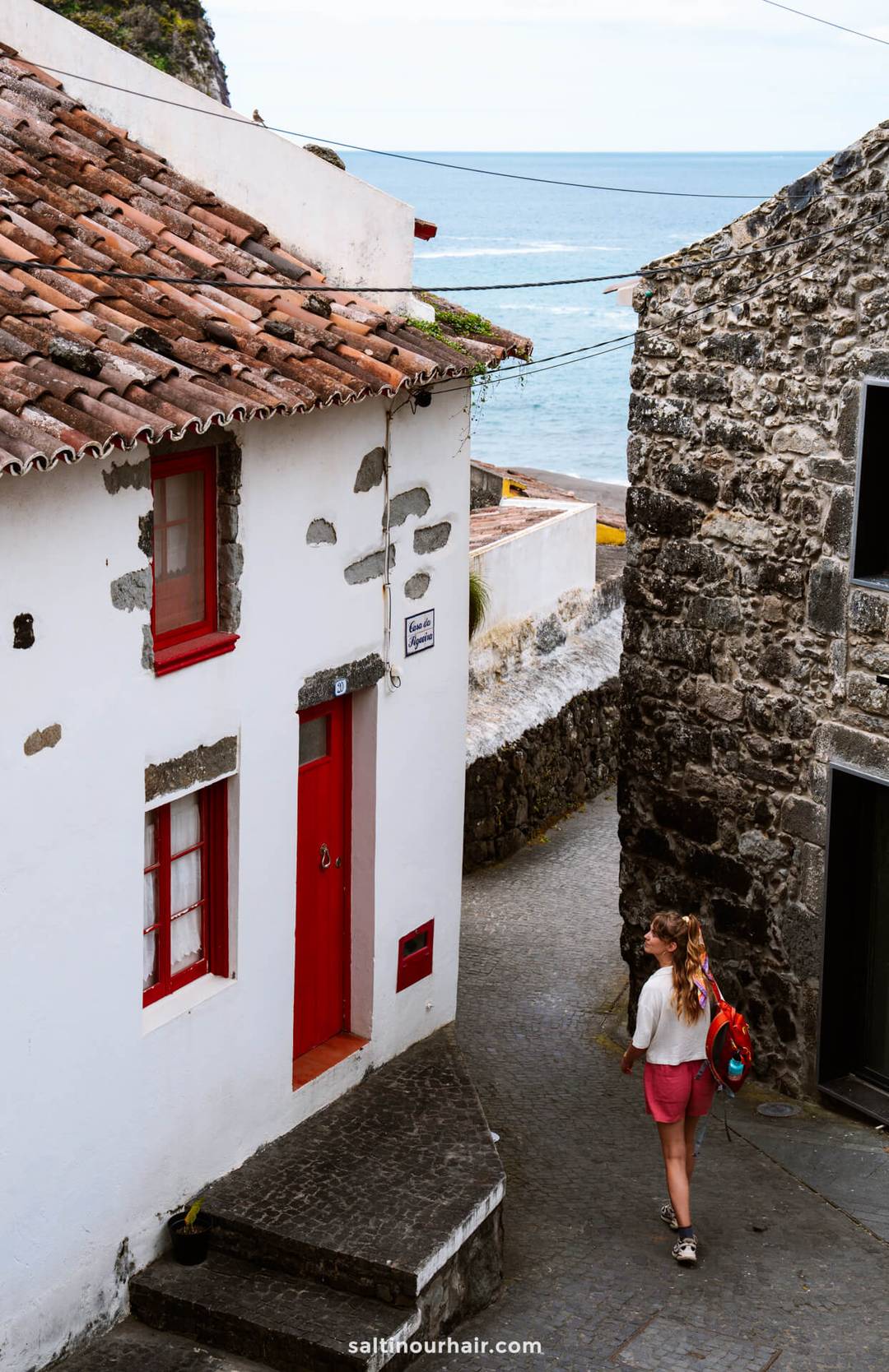 A woman with a backpack walks down a narrow cobblestone street lined with stone buildings near the ocean, exploring one of the charming things to do in S&atilde;o Miguel, Azores.