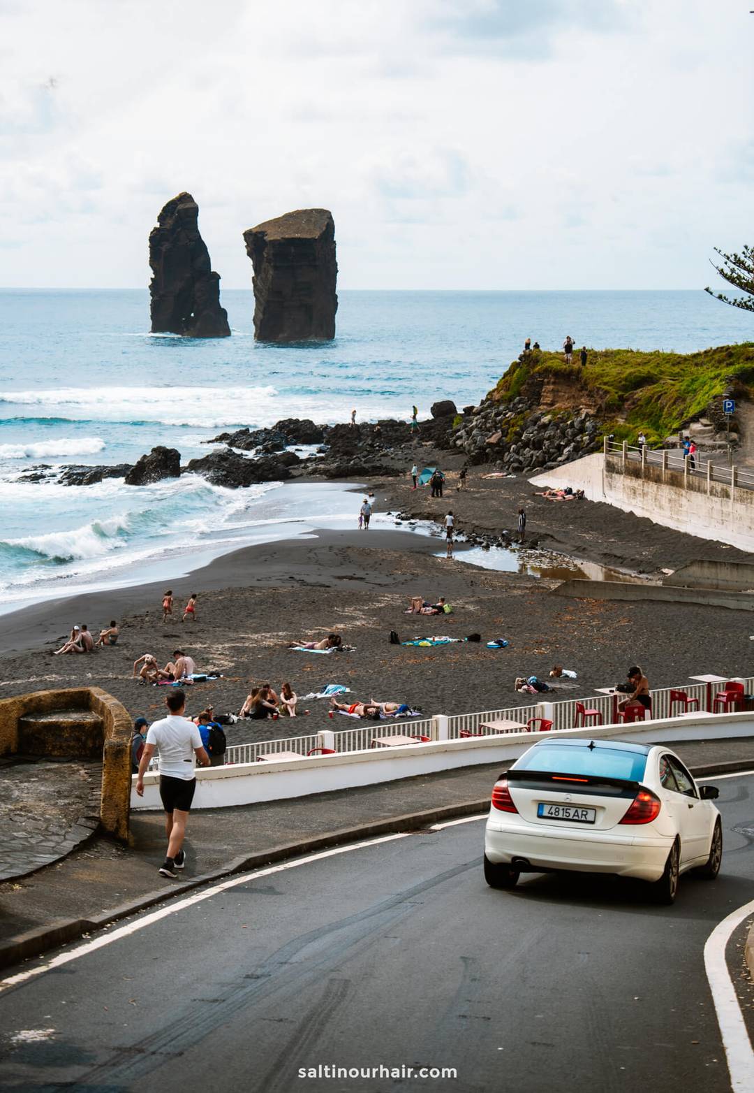 People relax on a black sand beach near the ocean, with two large rock formations in the water&mdash;a must-see among things to do in S&atilde;o Miguel. A car drives down a road in the foreground.