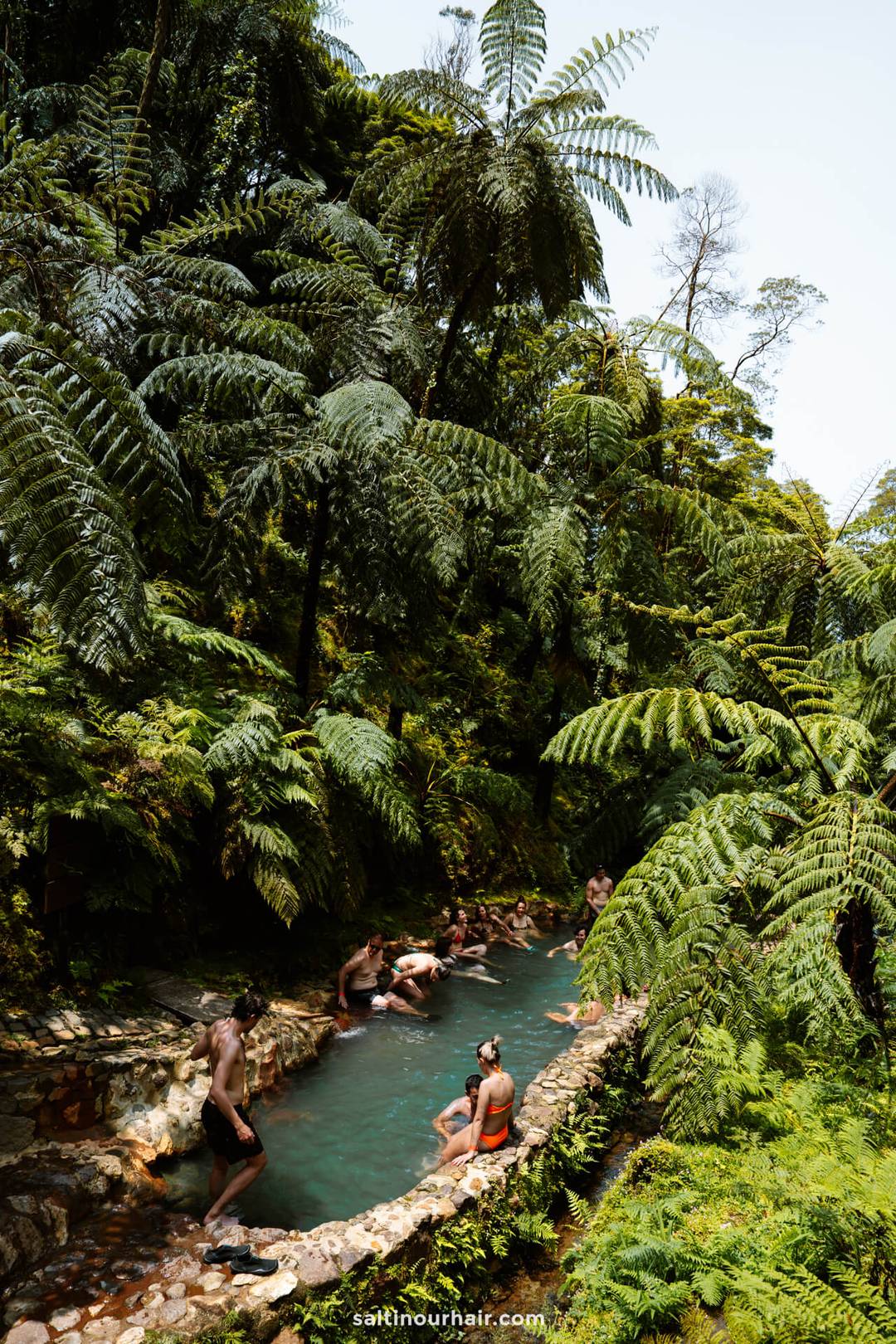 A group of people relax in a narrow, natural hot spring pool surrounded by dense, green tropical vegetation and tall ferns&mdash;one of the unforgettable things to do in S&atilde;o Miguel, Azores.