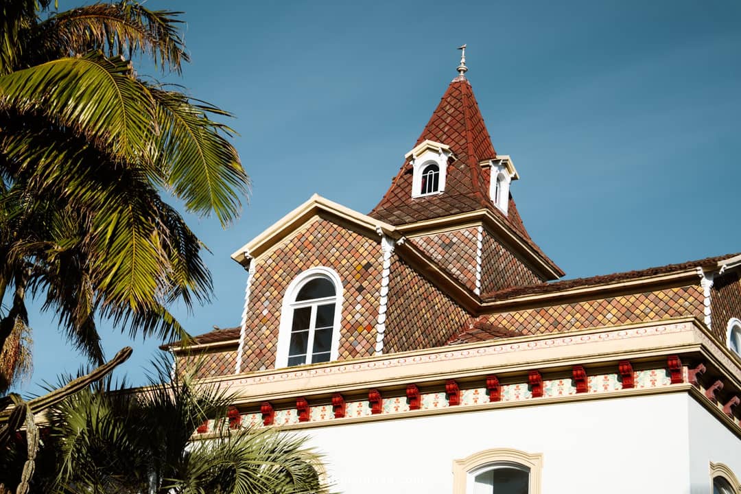A building with a red-tiled roof and tower, white walls, and decorative trim, set against a clear blue sky with palm trees in the foreground&mdash;one of the charming sights among the many things to do in S&atilde;o Miguel.