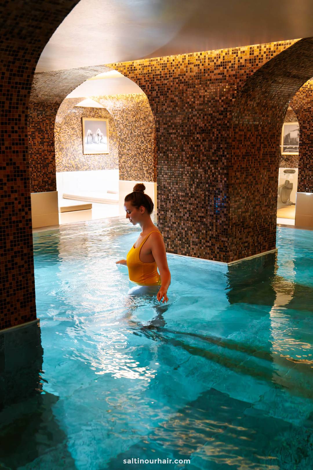 A woman in a yellow swimsuit walks in an indoor pool with mosaic tile walls and arched ceilings, experiencing one of the unique things to do in S&atilde;o Miguel, Azores.