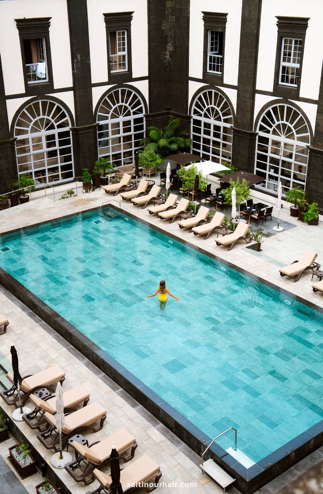 A woman in a yellow swimsuit stands in the center of a rectangular outdoor pool&mdash;one of the relaxing things to do in S&atilde;o Miguel, Azores&mdash;surrounded by lounge chairs and a building with arched windows.