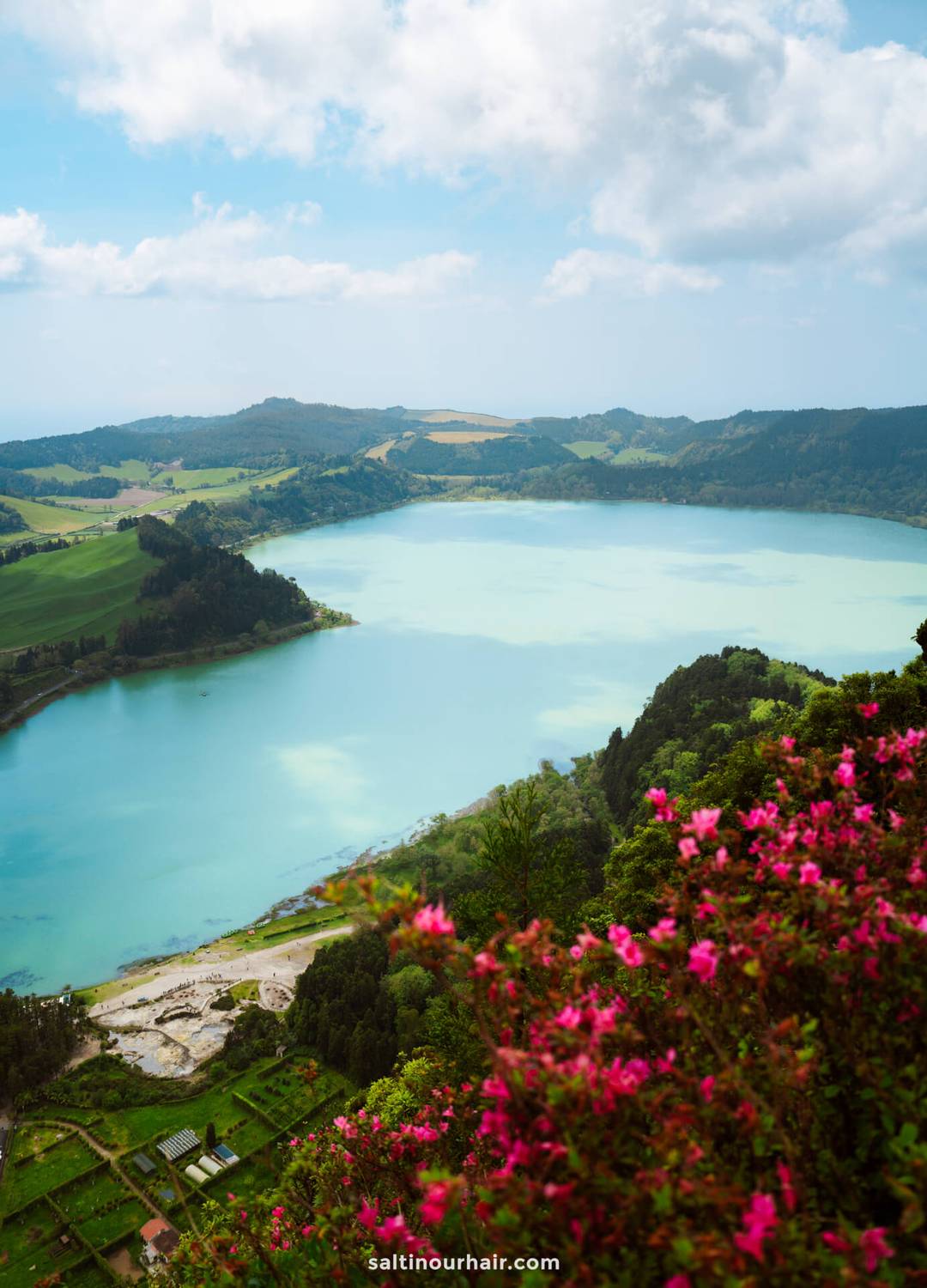 View of a turquoise lake called Lagoa das Furnas surrounded by green hills and fields, with pink flowers in the foreground under a partly cloudy sky&mdash;a perfect spot to add to your list of things to do in S&atilde;o Miguel, Azores.