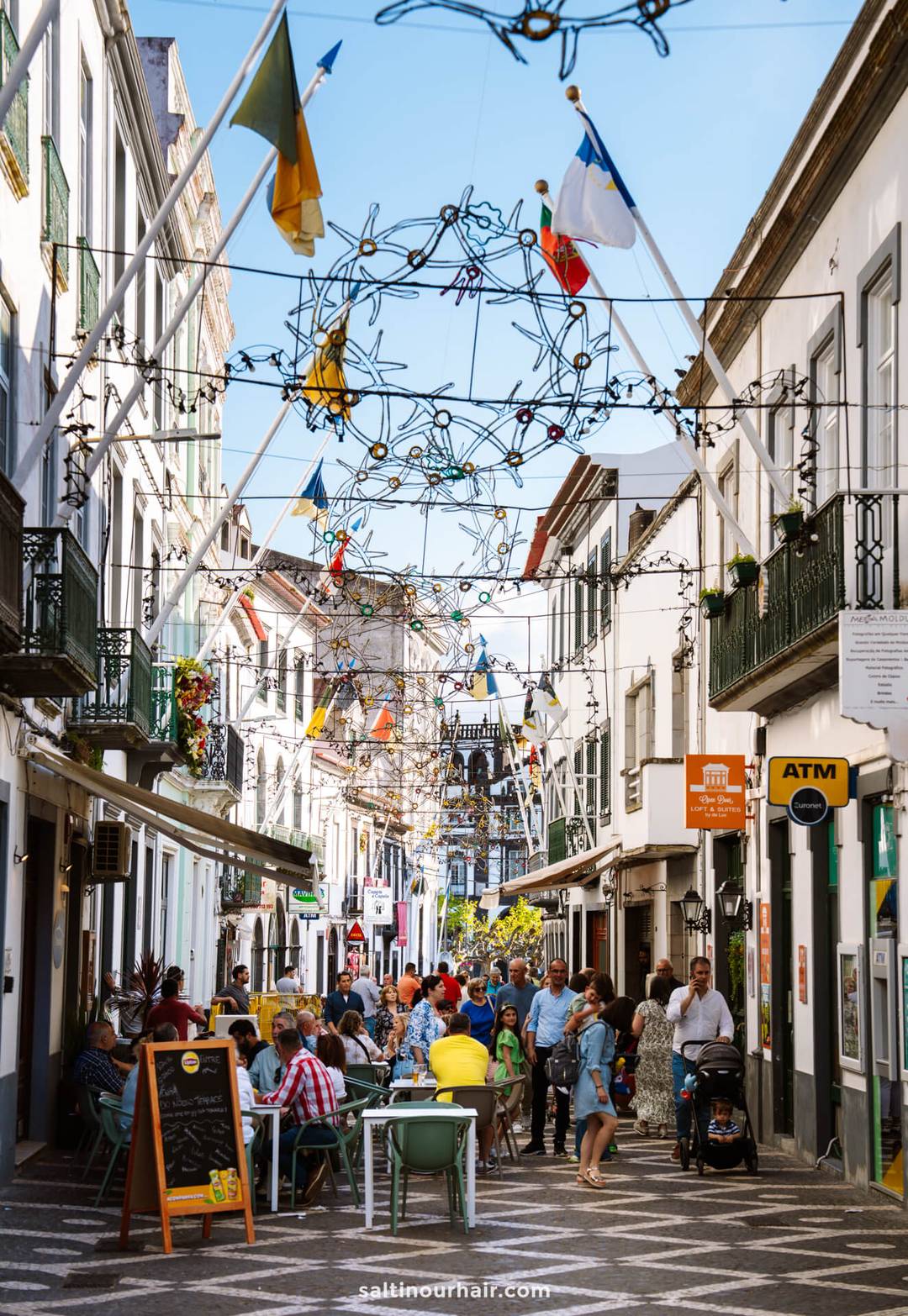 A lively pedestrian street in Ponta Delgada with outdoor caf&eacute; seating, people walking and dining, festive hanging decorations, various storefronts including an ATM&mdash;perfect for exploring local life and discovering things to do in S&atilde;o Miguel.