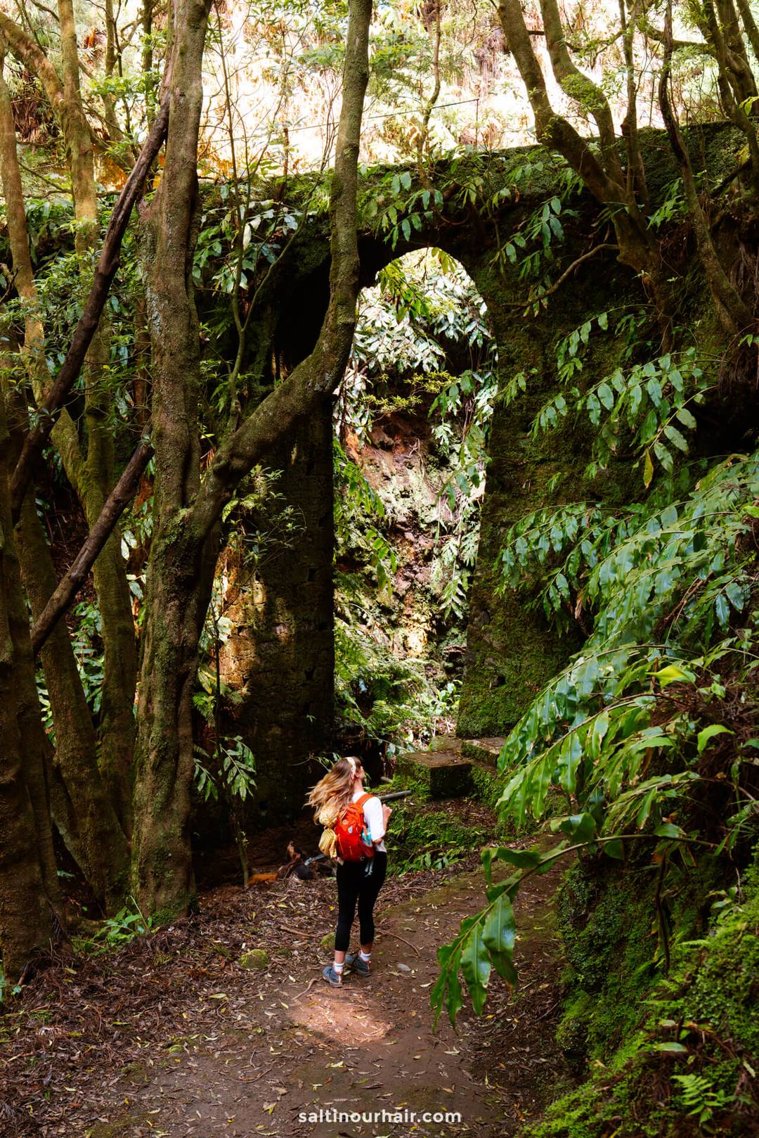 A woman with a red backpack stands on a forest path near a large, moss-covered stone arch surrounded by dense greenery on the PRC37 Janela do Inferno Hike&mdash;one of the unforgettable things to do in S&atilde;o Miguel, Azores for nature lovers and adventurers.