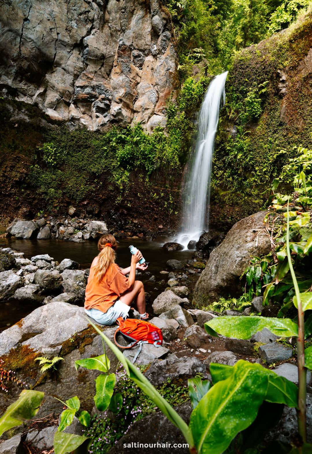 A person with long hair sits on rocks near a waterfall and pool, holding a camera&mdash;one of the top things to do in Furnas&mdash;surrounded by lush greenery and a backpack.