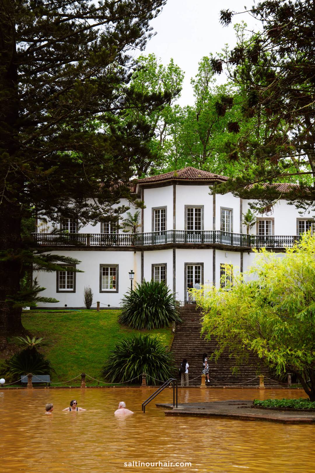 People swim in the large brownish thermal pool at Parque Terra Nostra hot springs, surrounded by lush greenery and a white two-story building in S&atilde;o Miguel, Azores.