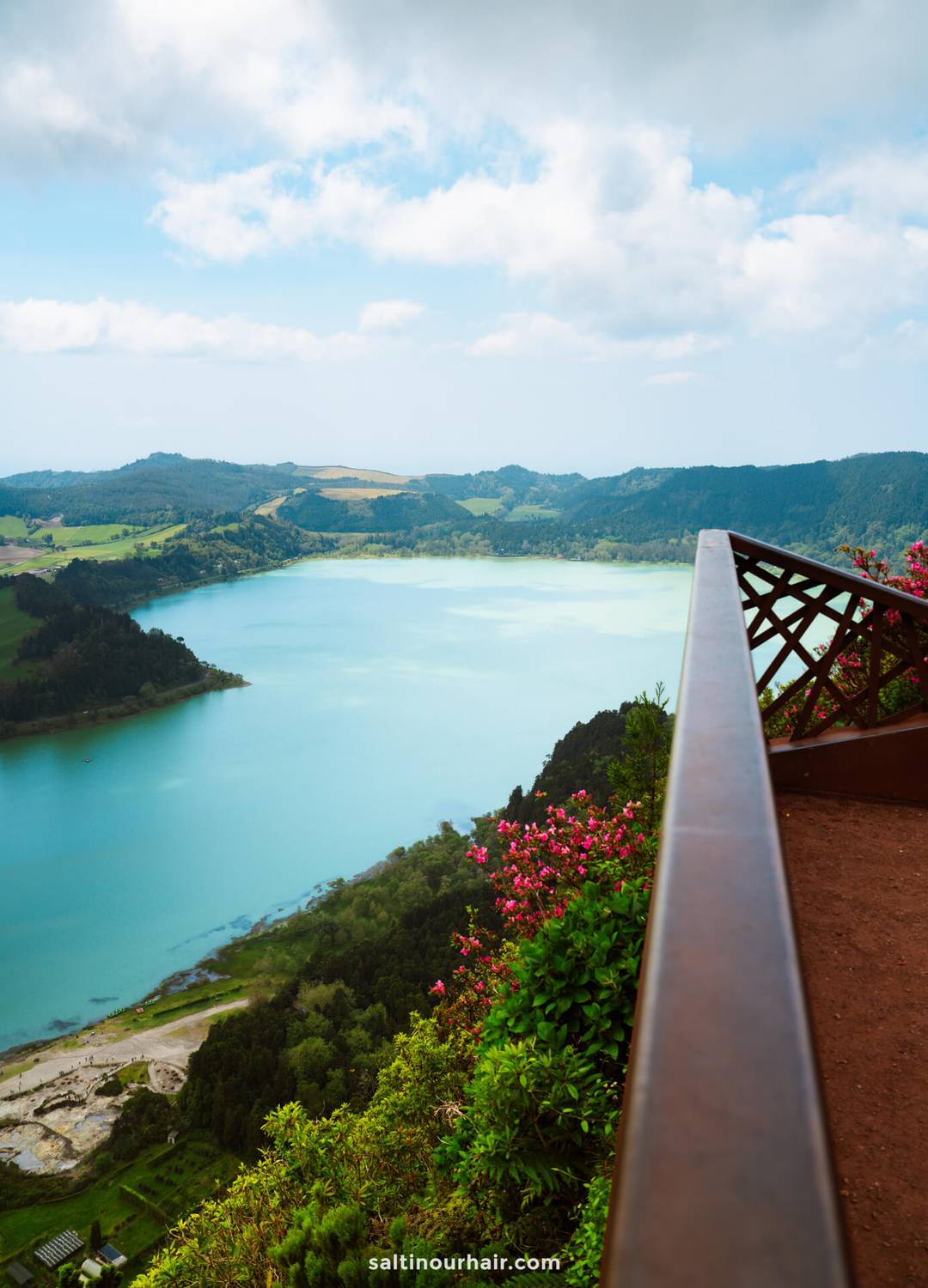 A scenic view of a turquoise lake from a viewpoint surrounded by lush green hills, seen from a wooden lookout with flowers in the foreground&mdash;what you can do when you rent a car azores