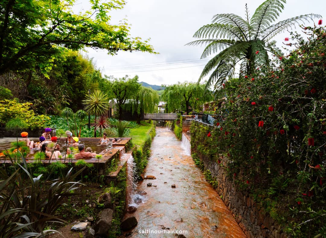 A group of people sit in a small outdoor hot spring pool at Po&ccedil;a da Dona Beija hot springs, beside a narrow, orange-colored stream surrounded by lush greenery and flowering plants in S&atilde;o Miguel, Azores.