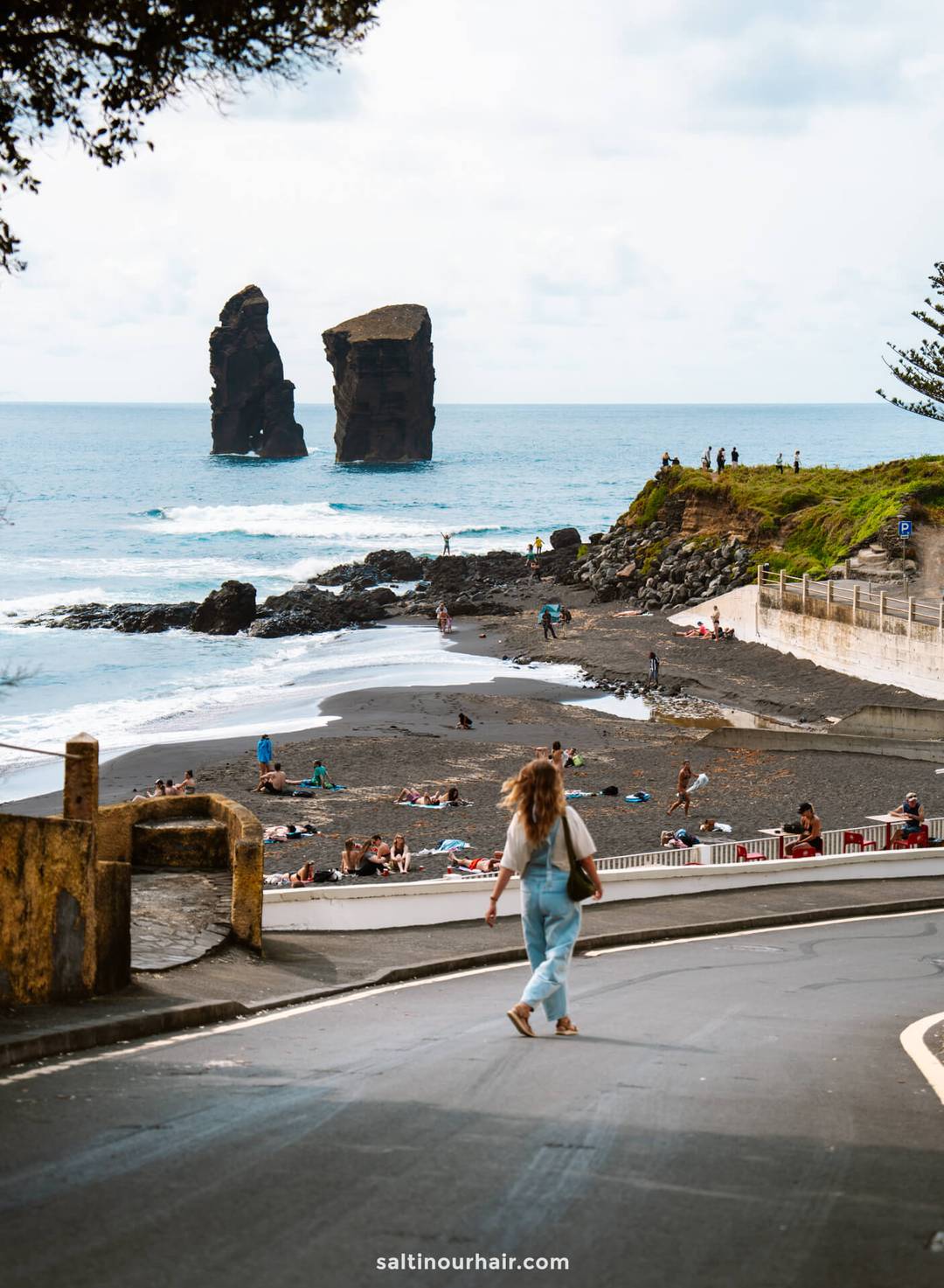 A woman crosses a road toward a black sand beach&mdash;an essential stop on any S&atilde;o Miguel itinerary&mdash;with people sunbathing and swimming; two large sea stacks rise from the ocean in the background.