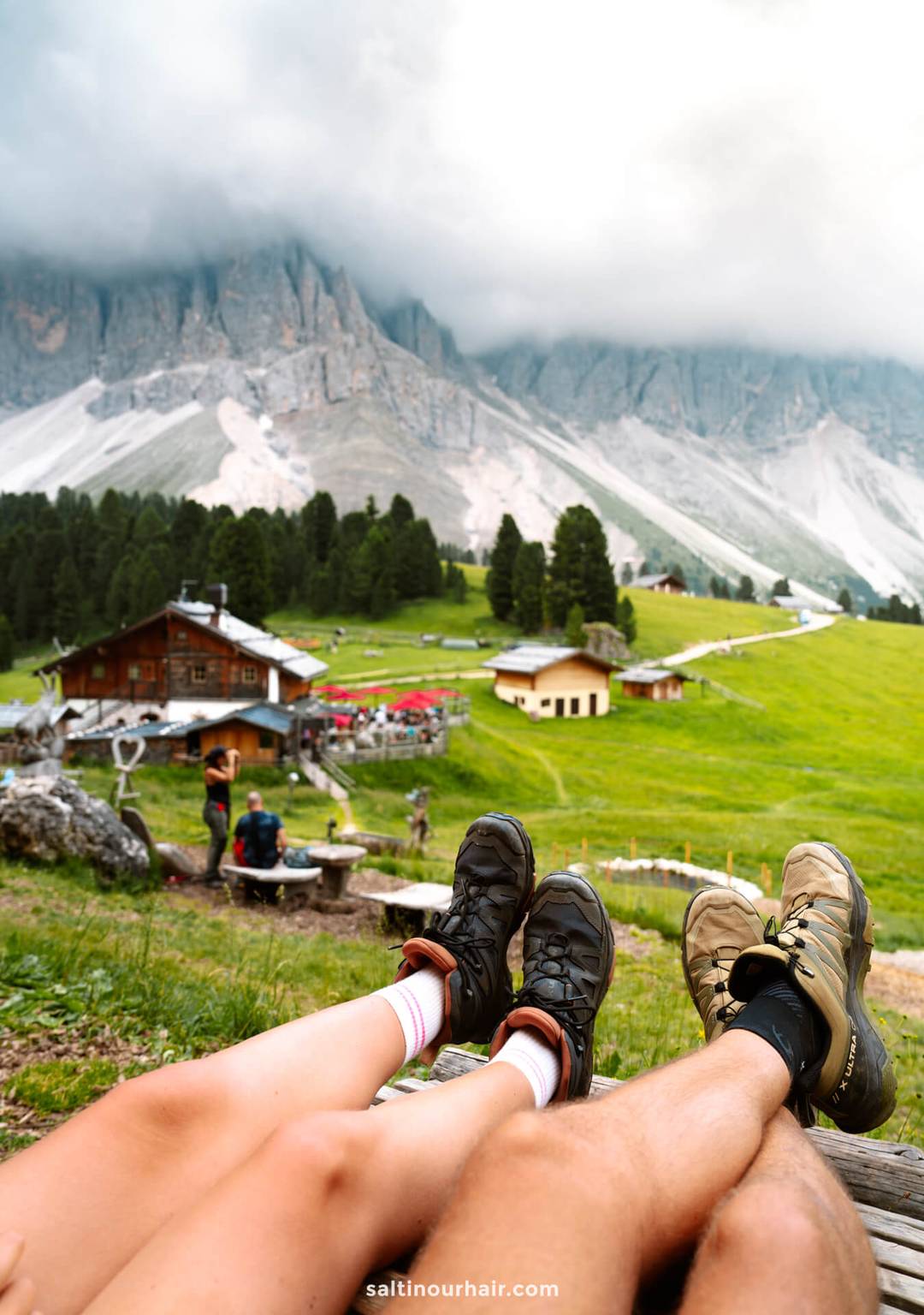 Two people with their legs outstretched, wearing hiking shoes, sit on grass overlooking alpine cabins and green hills during the Geisler Alm hike in the Dolomites, with stunning mountains and drifting clouds in the background.
