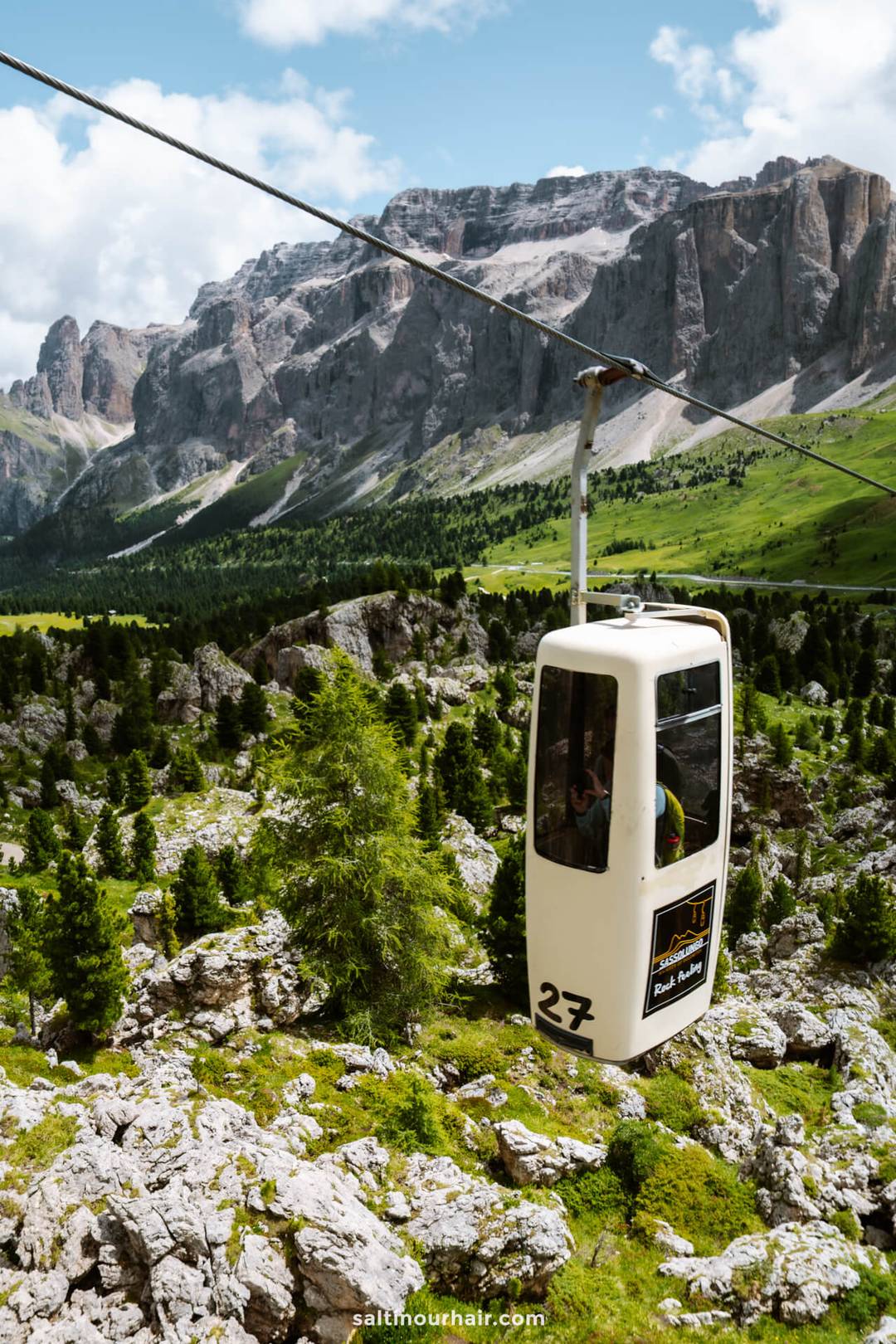 Een witte kabelbaan met nummer 27 glijdt over rotsachtig, groen alpenlandschap met hoge bergen op de achtergrond onder een gedeeltelijk bewolkte lucht, een van de leukste dingen om te doen in de Dolomieten.