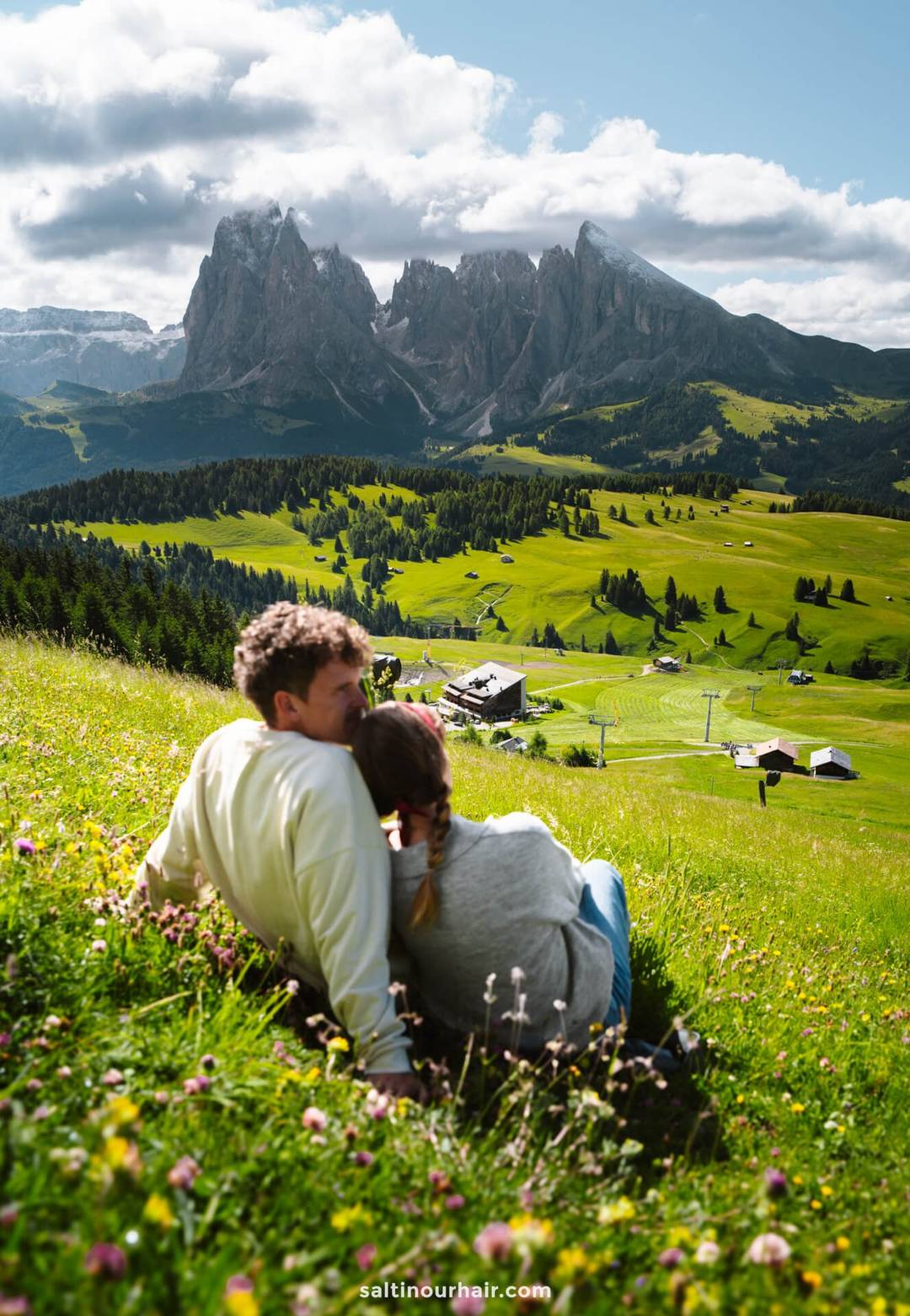 A couple sits on a grassy hillside overlooking a valley of houses and mountains in the background, enjoying the scenic beauty of Alpe di Siusi in the Dolomites, Italy, under a partly cloudy sky.