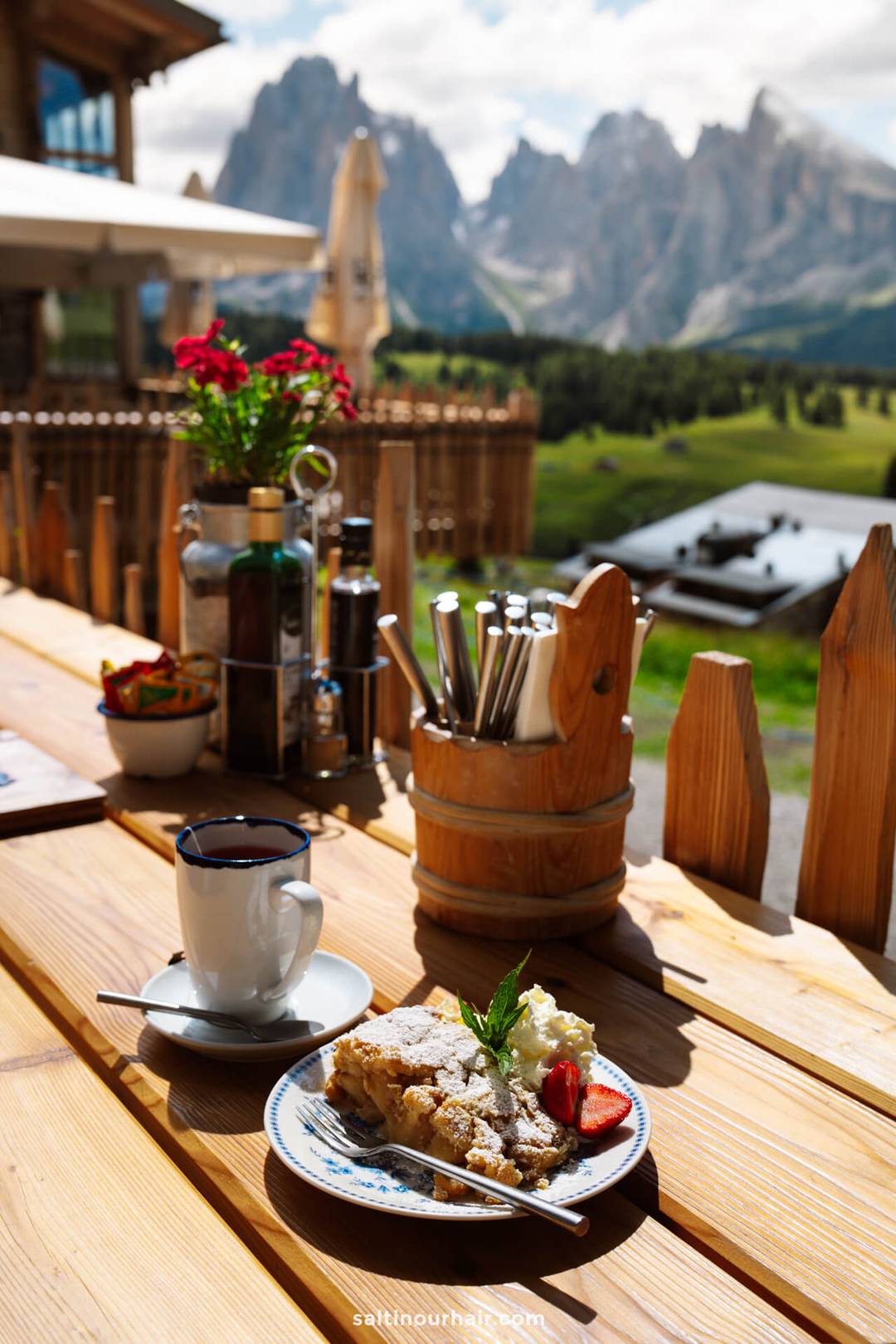 A slice of apfelstrudel with whipped cream and a strawberry, and a cup of coffee, rest on a wooden table outdoors with the stunning Seiser Alm Dolomites, Italy, providing a breathtaking mountain backdrop.
