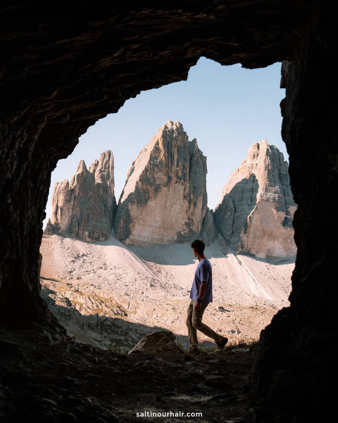 A person stands at the entrance of a rocky cave, looking at three large mountain peaks under a clear sky-one of the many unforgettable things to do in the Dolomites.