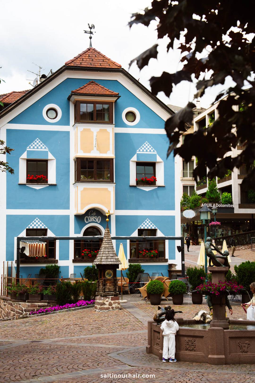 Een blauw-wit gebouw met bloembakken en gestreepte luifels aan een kasseienplein; een kind kijkt naar een bronzen standbeeld op de voorgrond, een charmant tafereel in de Dolomieten.
