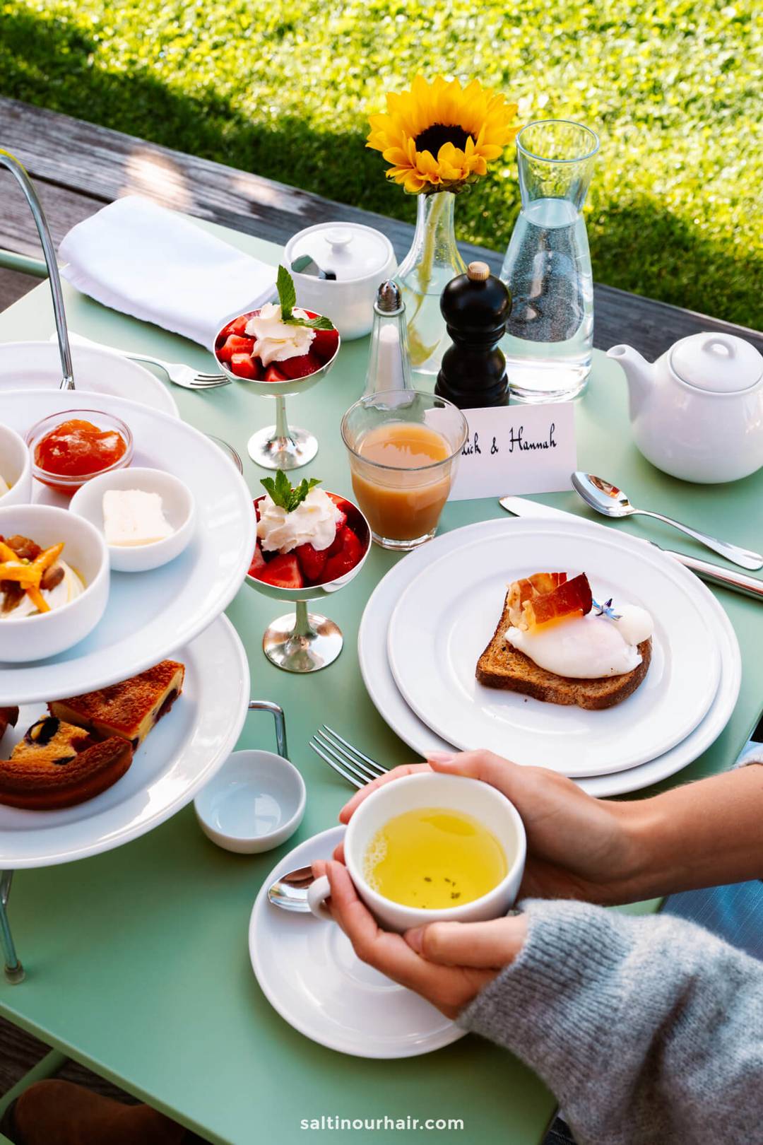A person enjoys breakfast at a hotel near Seiser Alm Dolomites, Italy, holding a cup of tea at an outdoor table set with pastries, jam, fruit parfaits, poached egg toast, sauces, a teapot, and water with a sunflower.