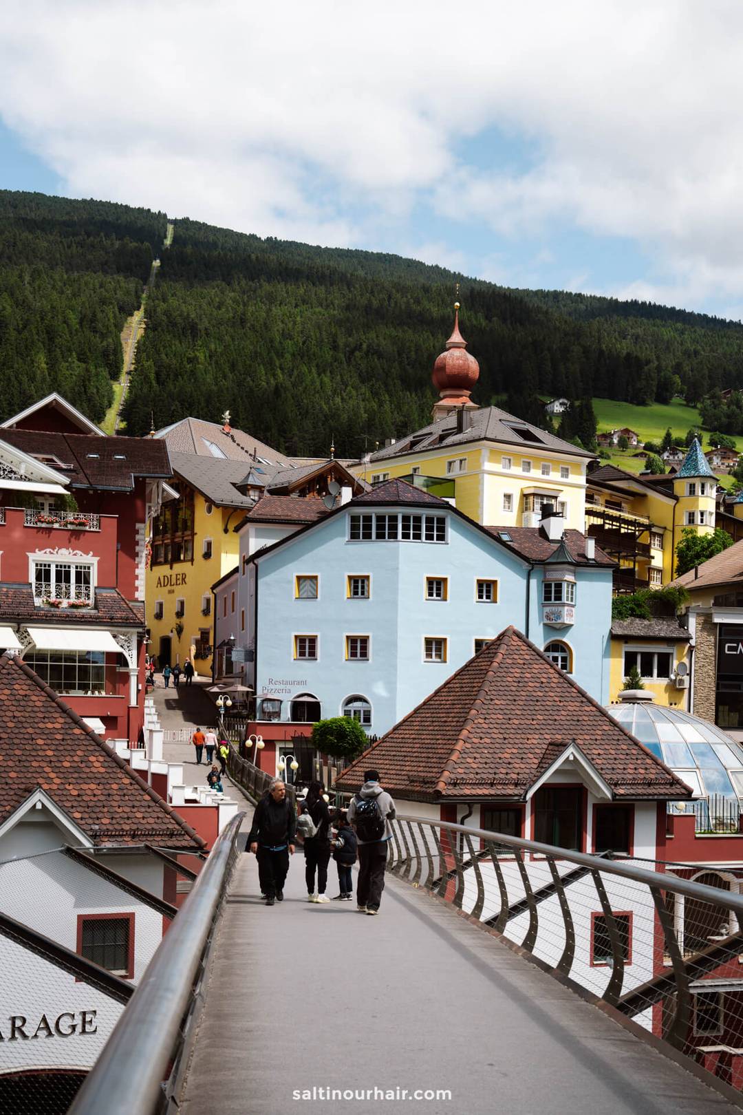 A group of people walks on a footbridge toward colorful buildings in Ortisei town surrounded by green hills and forests&mdash;one of the many charming places in the Dolomites italy