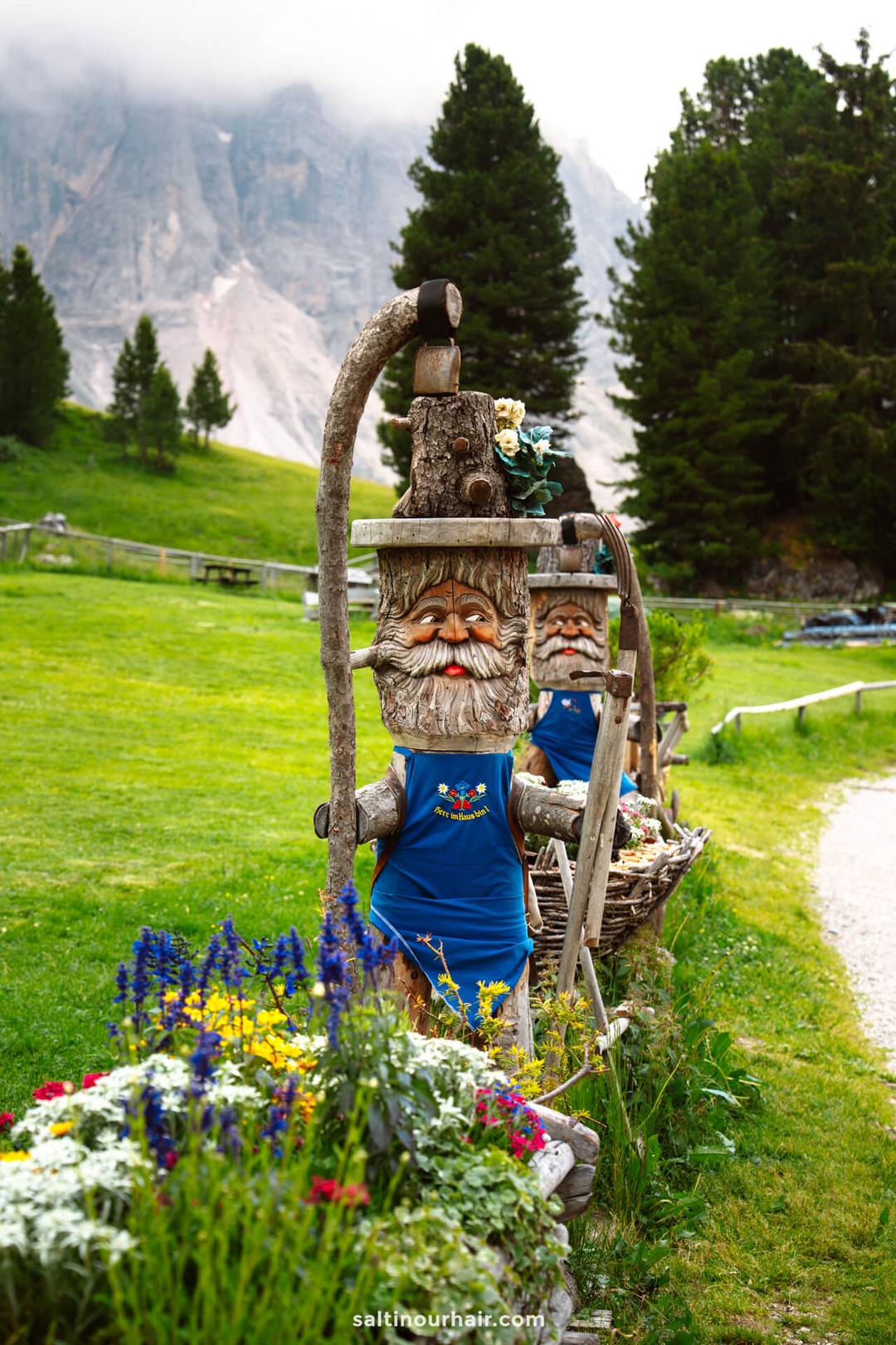Houten kabouter-beelden in blauwe schorten staan langs een pad in een grasrijk berglandschap met bloemen, een van de charmante dingen om te doen in de Dolomieten.