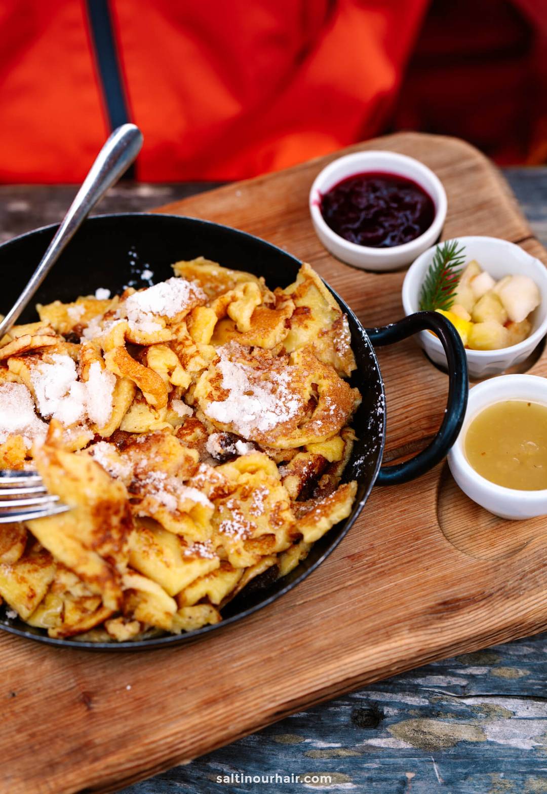 Een koekenpan met Kaiserschmarrn bestrooid met poedersuiker, geserveerd op een houten plank met vruchtencompote, stukjes fruit en saus, een authentieke traktatie in de Dolomieten.