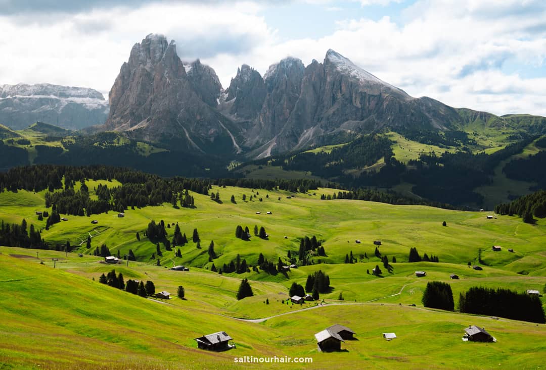 Green rolling hills dotted with small wooden cabins and trees stretch across the Seiser Alm Dolomites, Italy, with rugged mountains rising in the background under a partly cloudy sky.