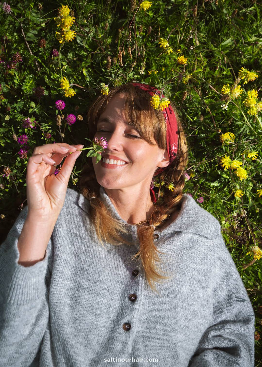A woman with braided hair and a red bandana lies on grass with wildflowers in Alpe di Siusi, Dolomites, Italy, smiling and holding a small flower near her nose.