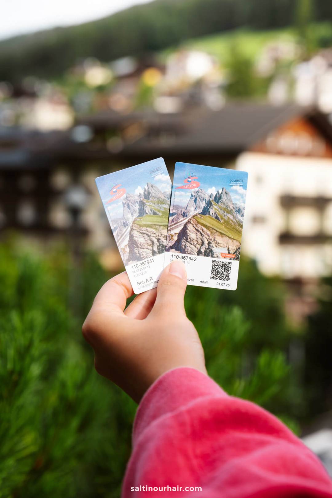 Een hand houdt twee schilderachtige bergkabelbaan tickets vast met een onscherpe achtergrond van gebouwen en bomen&mdash;een van de must-try dingen om te doen in de Dolomieten.
