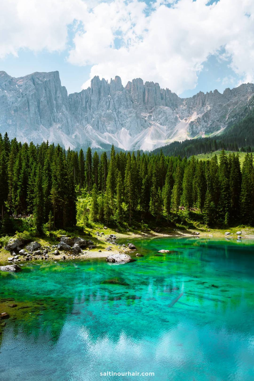 Clear turquoise lake called lago di carezza surrounded by pine trees and rocky mountains under a partly cloudy sky&mdash;a perfect spot to add to your list of things to do in the Dolomites.