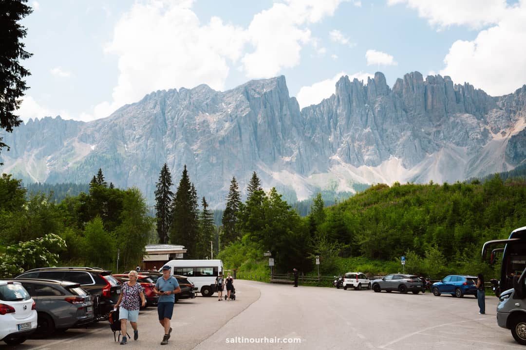 A parking lot with several cars and people, surrounded by green trees, with tall jagged mountains in the background under a partly cloudy sky&mdash;an ideal starting point for exploring things to do in the Dolomites.