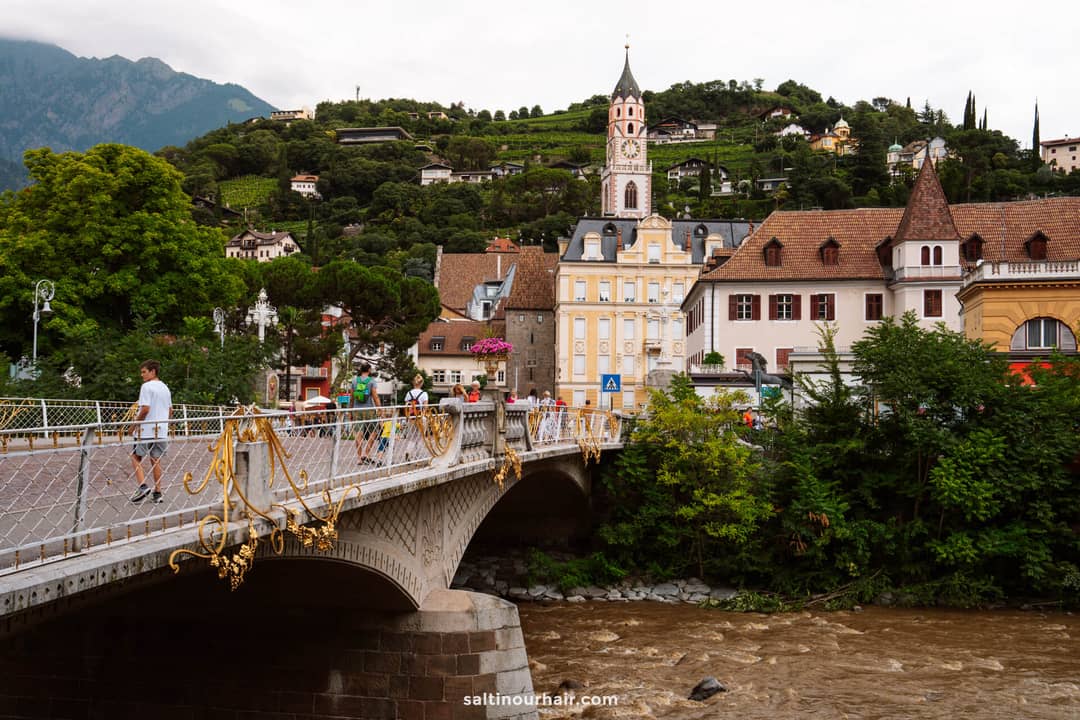 A pedestrian bridge with ornate railings crosses a brown river, leading to historic buildings and a clock tower set against a green, hilly backdrop&mdash;an inviting scene among the many things to do in the Dolomites.