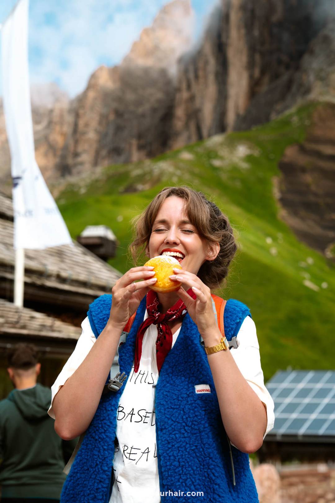 Vrouw staat buiten in een bergachtig gebied, glimlachend terwijl ze een broodje eet, een welverdiende pauze tijdens het verkennen van de Dolomieten. Ze draagt een blauw vest, rode sjaal en een wit shirt met tekst.