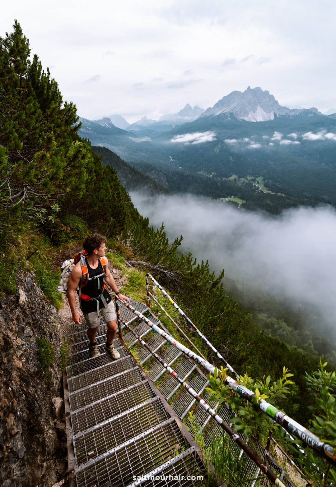A hiker with a backpack climbs a metal staircase on a mountain trail to lago di sorapis in the Dolomites, surrounded by trees, mist, and distant peaks&mdash;a stunning adventure and one of the top things to do in the Dolomites italy