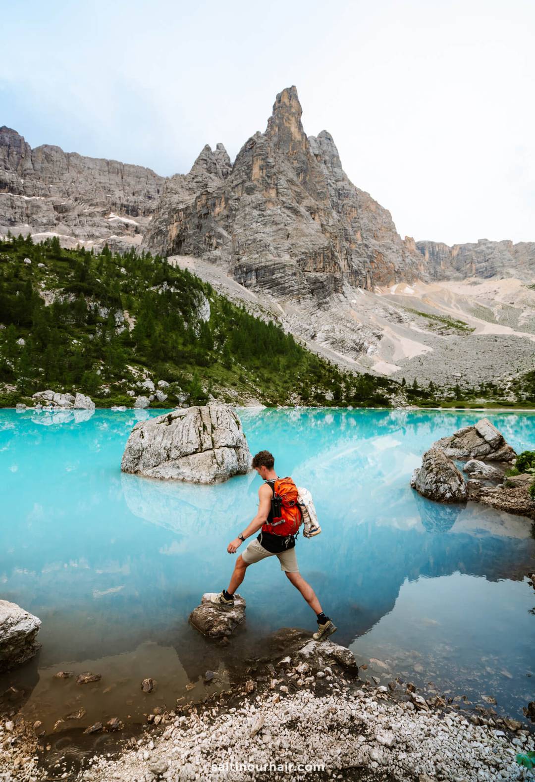 A man with a red backpack steps on rocks by a turquoise mountain lake called lago di sorapis, surrounded by steep rocky peaks and green trees&mdash;one of the unforgettable things to do in the Dolomites italy