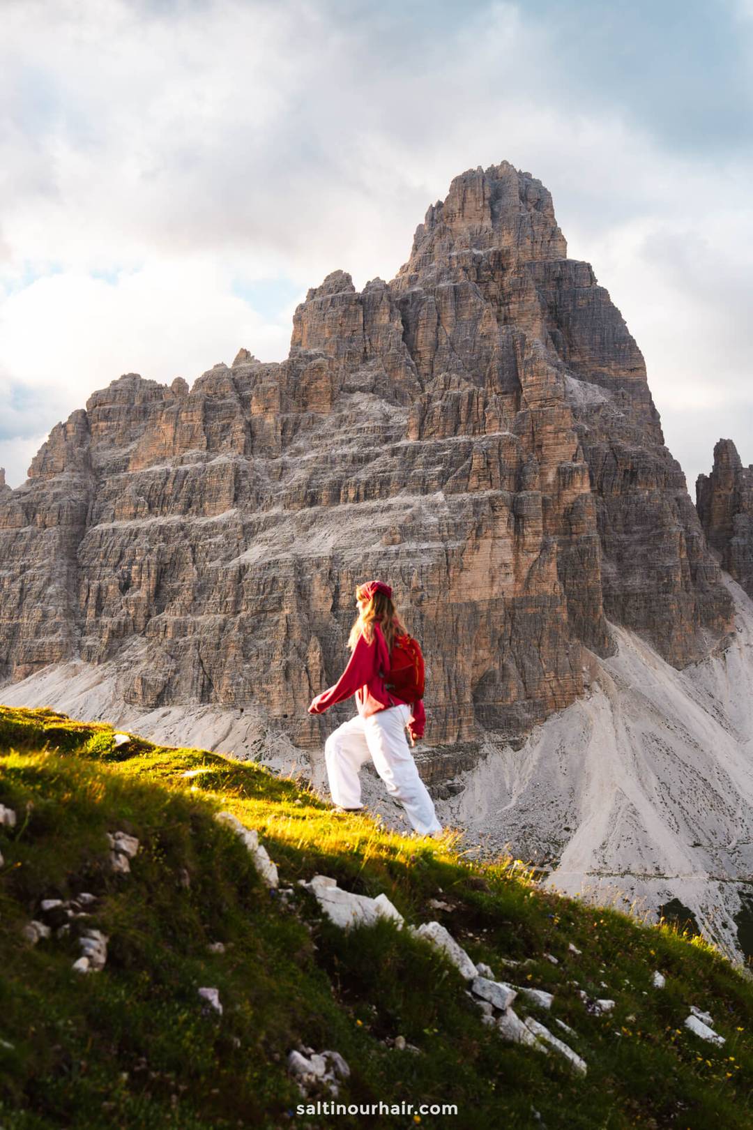 A woman wearing a red jacket and white pants hikes up a grassy slope with a large rocky mountain in the background&mdash;Cadini di Misurina is one of the unforgettable things to do in the Dolomites.