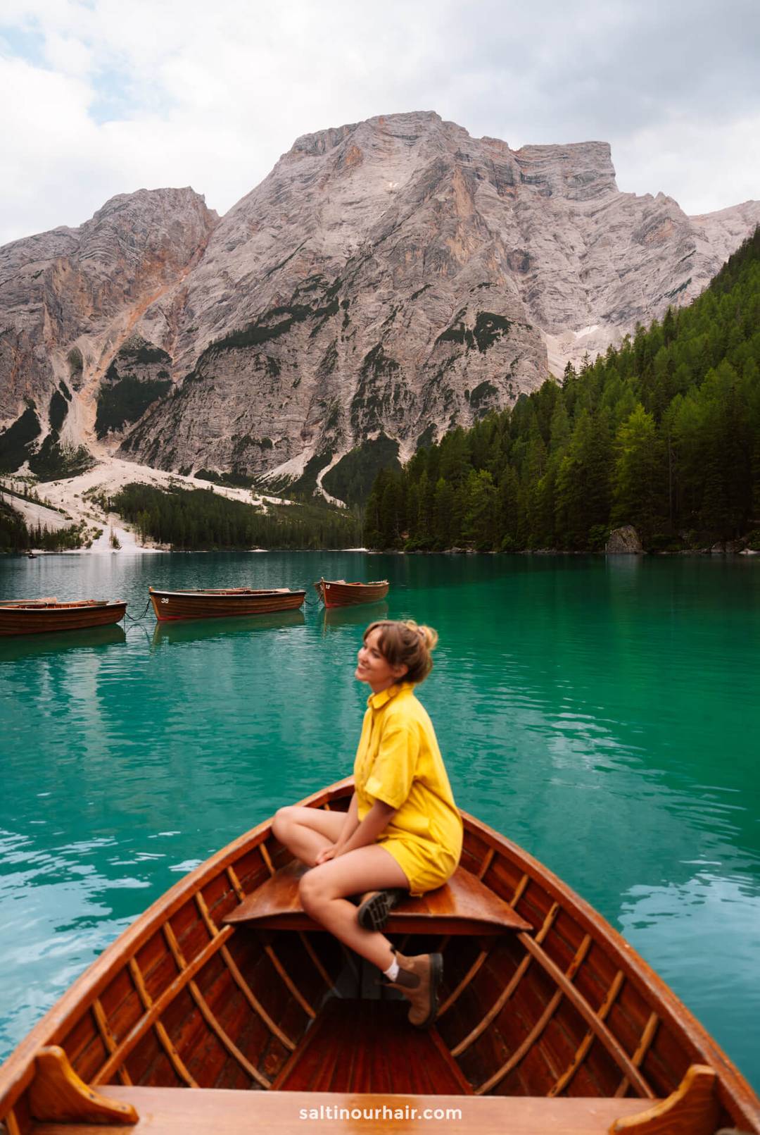 A woman in a yellow outfit sits at the front of a wooden boat on a turquoise lake, surrounded by mountains and pine trees-one of the unforgettable things to do in the Dolomites.