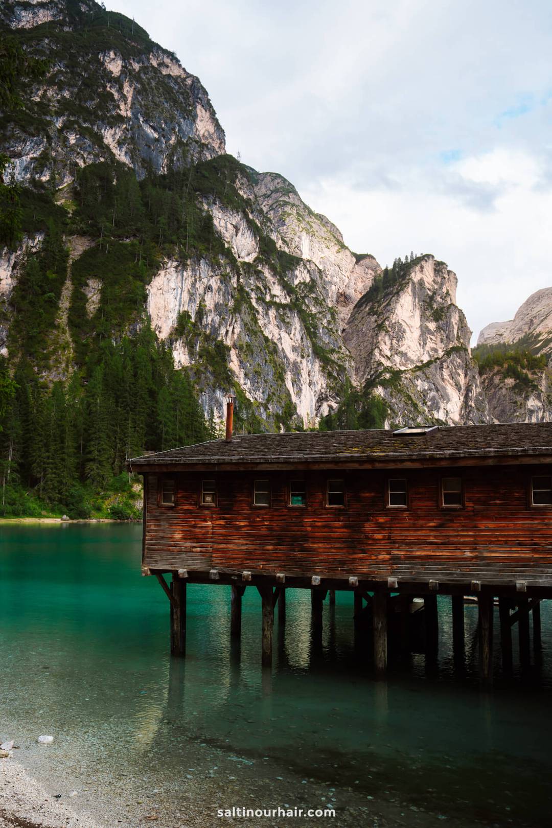 A wooden boathouse on stilts over a turquoise lake, surrounded by forested mountains and rocky cliffs under a partly cloudy sky-one of the most picturesque things to do in the Dolomites.