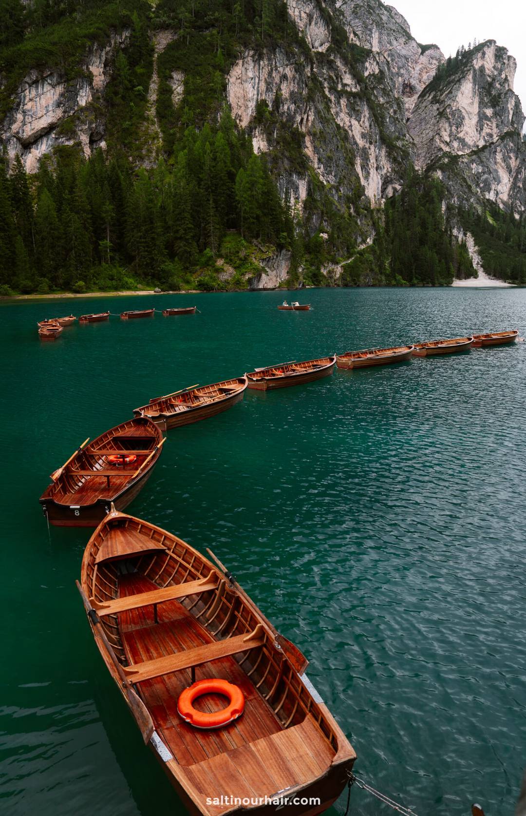 Wooden rowboats are lined up in a curved formation on a turquoise lake, framed by pine forest and rocky mountains under an overcast sky-an iconic scene among the unforgettable things to do in the Dolomites.