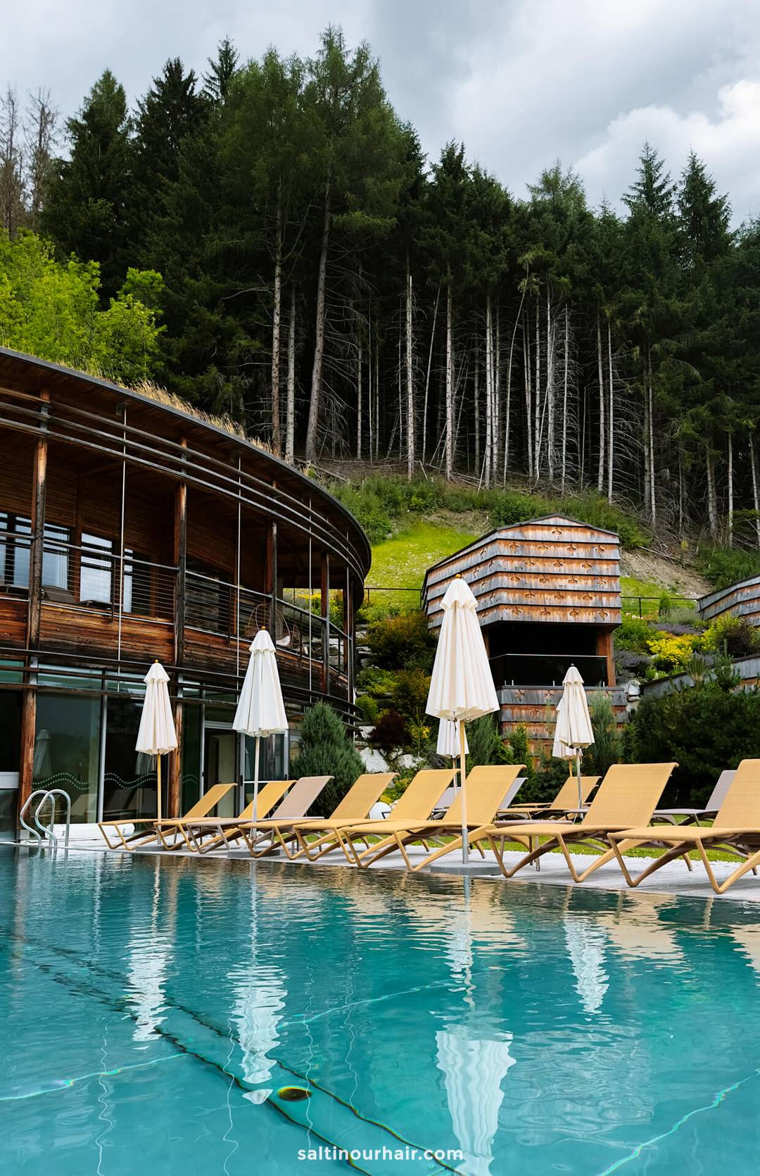 Outdoor pool with yellow lounge chairs and white umbrellas, modern wooden buildings, and a backdrop of dense forest under a cloudy sky&mdash;an idyllic spot for relaxing after exploring the top things to do in the Dolomites.