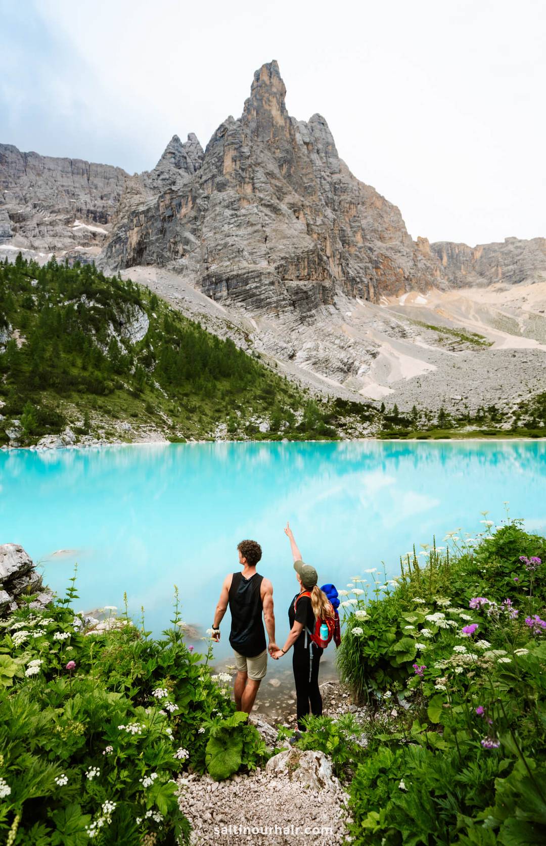 Stel houdt elkaars hand vast bij het turquoisblauwe Lago di Sorapis in de Dolomieten, Itali&euml;, terwijl &eacute;&eacute;n persoon wijst naar een hoge rotsachtige