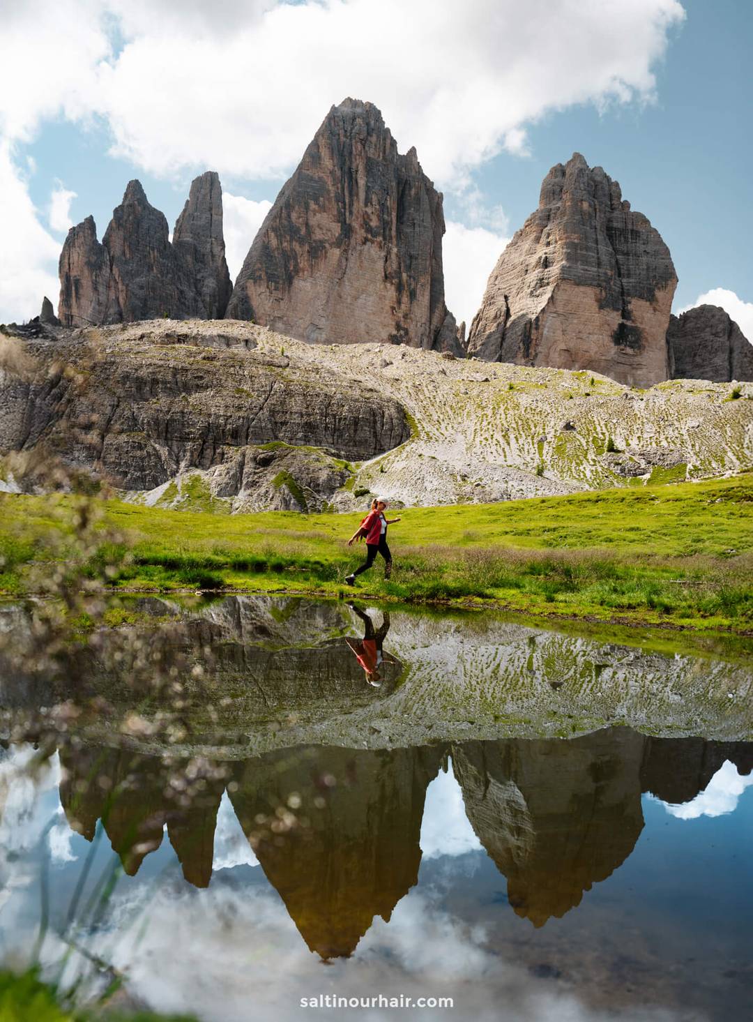 Person in a red jacket walks by a small pond reflecting the Tre Cime peaks and partly cloudy sky, set within a grassy, rocky landscape.