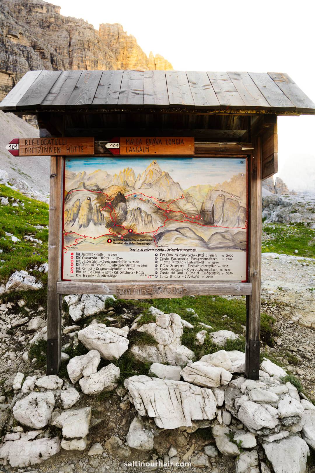Wooden trail signboard with a map and directions for hiking routes in the Tre Cime rocky mountain area, surrounded by grass and stone terrain.