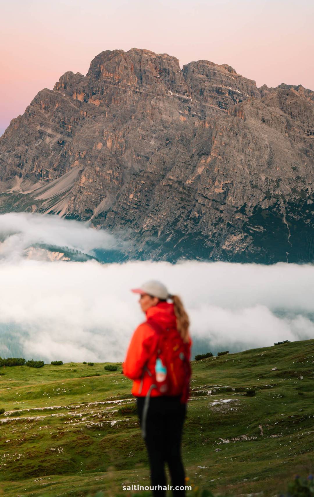 A person in a red jacket stands on a grassy field with Tre Cime&rsquo;s iconic peaks and low clouds in the background.