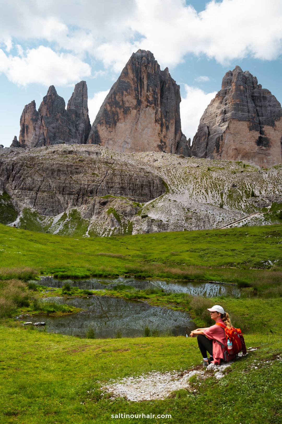 A person with a red backpack sits on grass near a small pond, with the iconic Tre Cime&rsquo;s rocky peaks rising in the background under a partly cloudy sky.