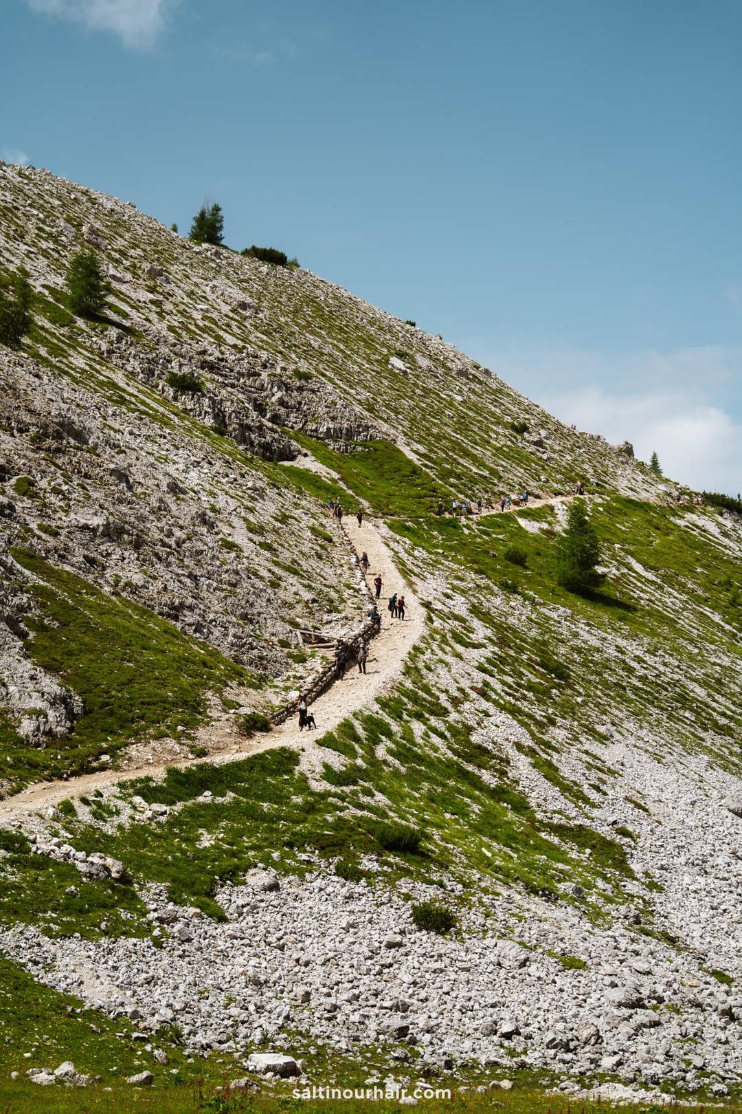 A group of hikers walk along a winding trail on a rocky, grassy hillside beneath the iconic peaks of Tre Cime and a partly cloudy sky.
