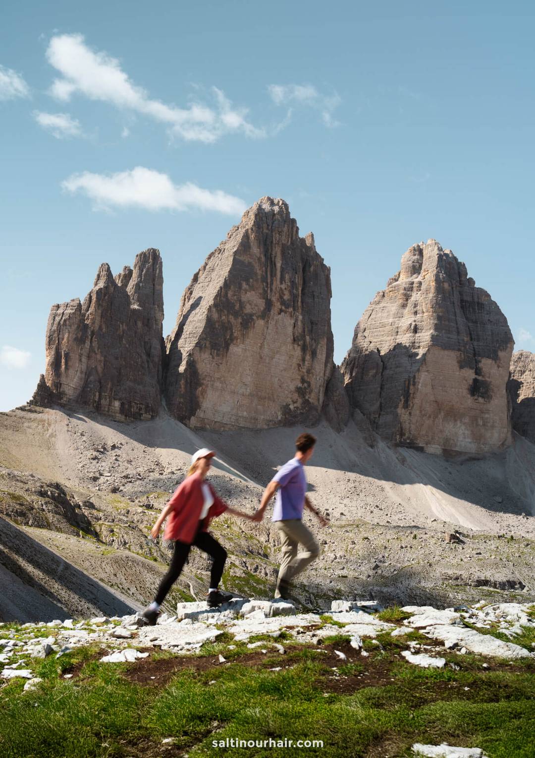 A couple walks in the foreground, framed by the majestic rocky peaks of Tre Cime rising in the background beneath a clear sky.