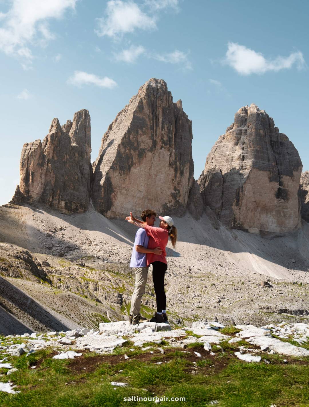 A couple stands on rocky terrain, with one person pointing towards the iconic Tre Cime peaks in the background beneath a partly cloudy sky.