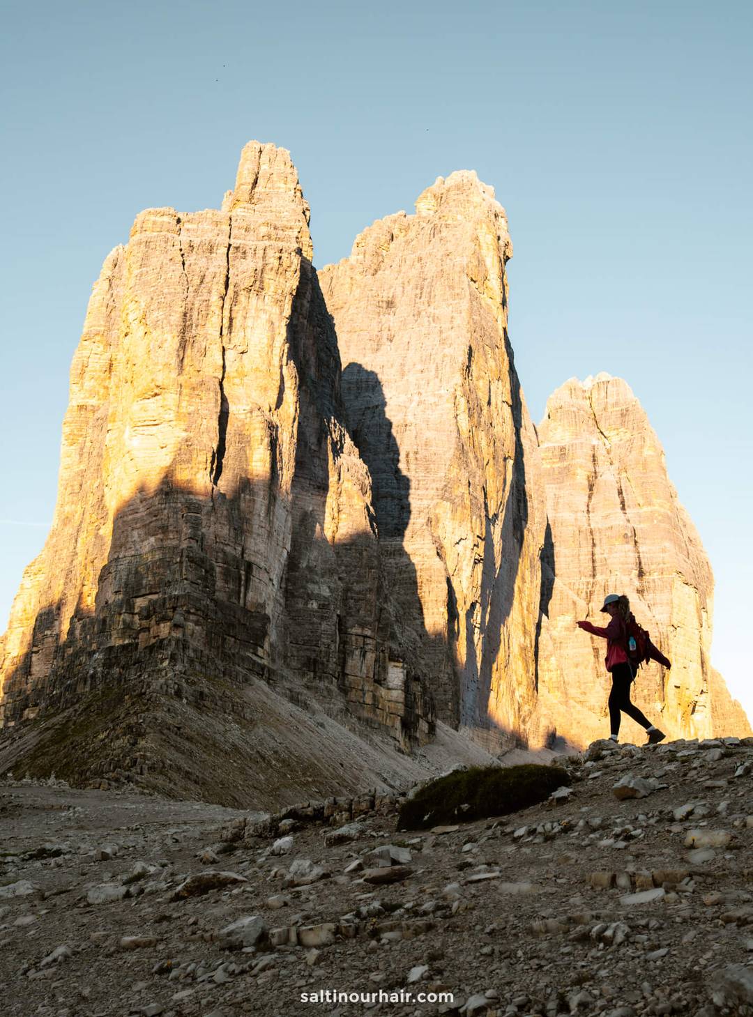 A person wearing a backpack stands on rocky terrain, pointing toward the sunlit peaks of Tre Cime under a clear sky.